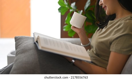 Calm Asian Woman Drinking Hot Tea And Reading Book On Comfortable Couch At Home