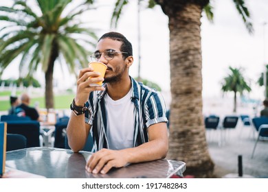 Calm Bearded Ethnic Man In Trendy Outfit And Eyeglasses Drinking Hot Beverage In Disposable Cup And Looking Away While Relaxing In Modern Outside Cafe