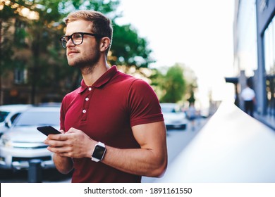 Ruhiges, bärtige Männchen in trendigem Outfit und Brille mit Smartphone in der Stadtstraße und Blick bei Tageslicht