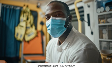 Calm Black African American EMS Professional Paramedic Looks At Camera While Wearing A Safety Face Mask In Ambulance Vehicle. Successful Emergency Medical Technician Outside The Healthcare Hospital.