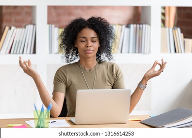 Calm black businesswoman taking break for meditation doing yoga exercise at workplace, happy mindful african female worker student feeling zen no stress free relief balance harmony relaxing at work