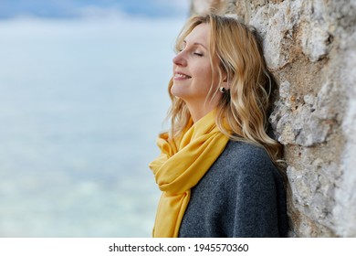Calm blonde woman with eyes closed yellow scarf and her hair blowing in the breeze stands near sea