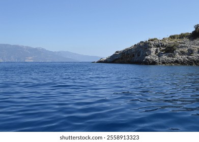 Calm blue waters of the Mediterranean meeting a rugged, rocky coastline with distant mountain views under a clear sky in Oludeniz, Turkey - Powered by Shutterstock