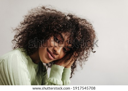 Calm brown-eyed woman in green top looks into camera. Charming dark-skinned brunette lady smiles gently on white background.
