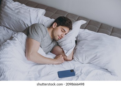Calm brunet tranquil young man in pajamas grey t-shirt lying in bed sleep near mobile cell phone slumber rest with closed eyes relax at home indoors bedroom. Good mood night morning bedtime concept.