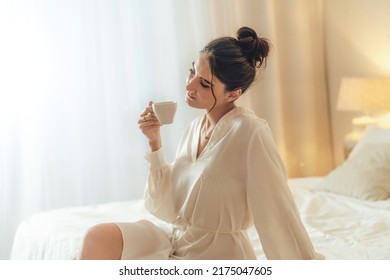 Calm Brunette Woman In White Silk Bathrobe Sitting On Bed And Drinking Coffee At The Sunny Morning Bedroom. Me Time Concept