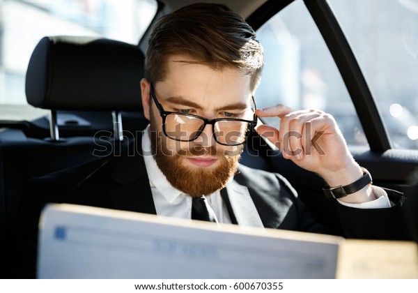Calm business man in eyeglasses reading\
newspaper while sitting in back seat of a\
car