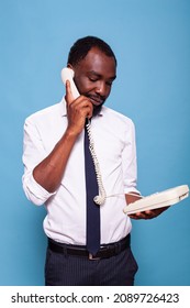 Calm Businessman Holding Reciever To Ear Standing Up Talking On Landline Cord Telephone Listening To Person Speaking At The Other End Of The Line. Employee In White Shirt Using Oldschool Phone.