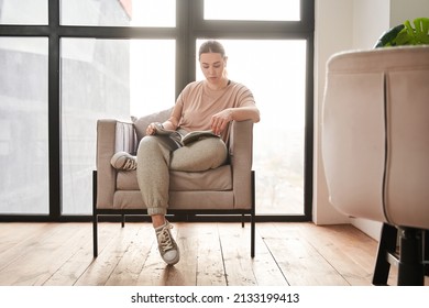 Calm Caucasian Female With Arm Implant Sitting At The Armchair And Reading Book