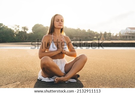 Calm Caucasian woman sitting in lotus pose enjoying free time for yoga practice at seashore beach, attractive female with closed eyes feeling good during zen research and asanas at coastline