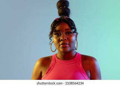 Calm, composed. Picture of young beautiful African girl, female model looking at camera isolated on blue green background in neon light. Concept of human emotion, facial expression. Copy space for ad.