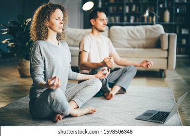 Ruhiges Paar in Pyjamas Meditieren, hören spirituelle Übungen auf Laptop, sitzen auf Lotus Pose zu Hause. Yoga-Konzept.