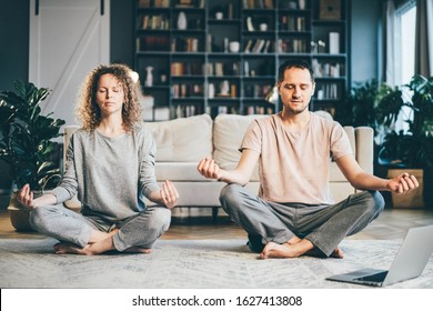 Calm Couple In Pajamas Meditating, Listening Spiritual Practices Lessons On Laptop, Sitting On Lotus Pose At Home. Yoga Concept.