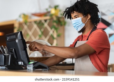 Calm Curly Attractive Young Black Man Bartender Or Waiter In Apron, Protective Mask Behind Bar Counter With Terminal Works On R-keeper In Cafe Interior. Small Business, Finance Due Covid-19 Pandemic