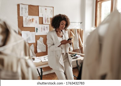 Calm curly brunette dark-skinned woman in white suit leans on desk in office of fashion designer and holds phone.