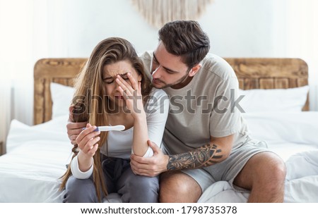 Calm down in difficult times and problems with conception. Sad man hugs crying woman and looks at pregnancy test in bedroom interior