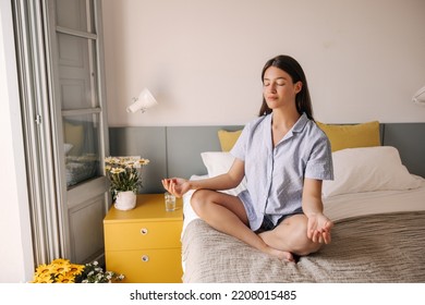 Calm down young caucasian girl with closing eyes meditating sitting on bed at home. Brunette hair woman dressed in cozy pajamas. Concept meditation, morning.