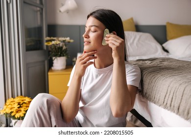 Calm Down Young Caucasian Woman Takes Care Of Her Face Sitting Opposite Window At Home. Brunette Hair Girl Wears In Pajamas. Concept Skin, Good Morning.