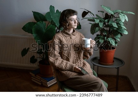 Calm dreamed woman wearing in vintage style in glasses with cup, smartphone in hands sitting on chair among plants at home looking at window. Pensive thoughtful young female resting relaxing alone.
