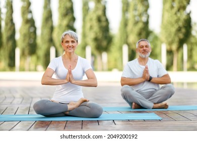 Calm elderly retired family couple practicing yoga outside happy husband and wife sitting on mats barefoot  in lotus position holding hands in namaste gesture and smiling at camera. Meditation and men