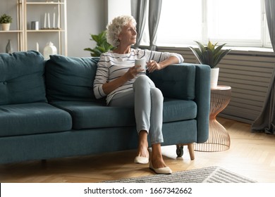 Calm Elderly Woman Sit On Comfortable Couch In Living Room Drink Tea Look In Window Distance Thinking Or Dreaming, Pensive Thoughtful Mature 50s Grandmother Enjoy Coffee Relax On Sofa At Home