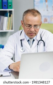 Calm Family Medical Doctor In Glasses Is Working On A Laptop Computer In A Health Clinic. Physician In White Lab Coat Is Browsing Medical History Behind A Desk In Hospital Office