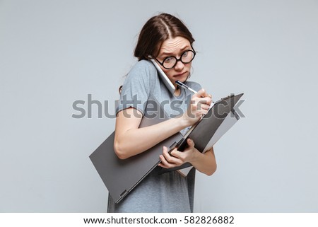 Calm female nerd in funny eyeglasses which talking on phone, holding laptop and notebook as well as writing something on clipboard