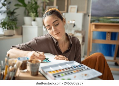 Calm female painter drawing with watercolor. Inspired pensive dreamy woman working on painting with focus immersed in creative process. Girl artist freely uses brushes, aquarelle to paint masterpieces - Powered by Shutterstock