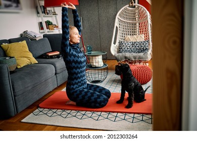 Calm, Flexible Senior Woman Kneeling On A Yoga Mat At Home And Doing Yoga With Her Dog.