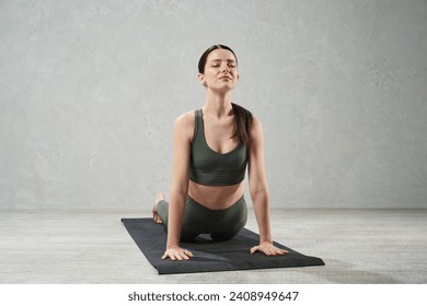 Calm focused woman in sports bra meditating in Cobra pose on gym mat. Front view of young inspired female practicing yoga in Bhujangasana with closed eyes, against grey background. Lifestyle concept.  - Powered by Shutterstock