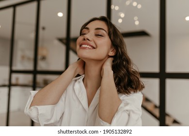 Calm Happy Brunette Woman In Cotton White Shirt Touches Neck And Smiles. Close Up Portrait Of Curly Lady In Light Blouse Poses At Home.