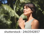 Calm happy young Hispanic woman holding hands in namaste meditating doing yoga breathing spiritual practice at retreat with eyes closed feeling peace of mind, standing in green nature tropical park.