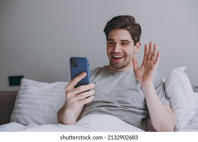 Calm Happy Young Man In Pajamas Grey T-shirt Lying Sit In Bed Talk By Video Call On Mobile Cell Phone Waving Hand Rest Relax At Home Indoors Bedroom. Good Mood Sleeping Night Morning Bedtime Concept.