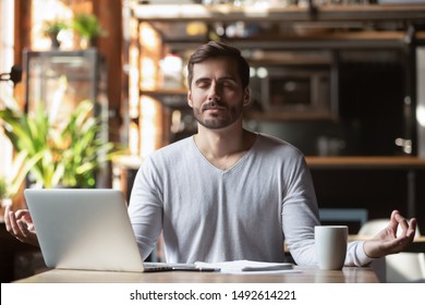 Calm healthy businessman meditate at work desk feeling zen no stress free relief, mindful young man taking break doing yoga exercise relax breath fresh air for peace of mind sit in office cafe