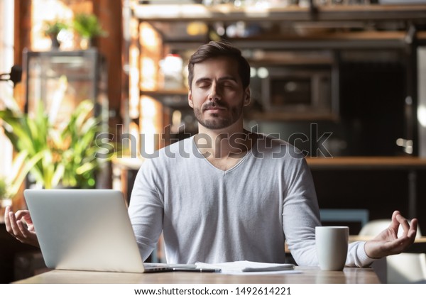 Calm healthy businessman meditate at work desk
feeling zen no stress free relief, mindful young man taking break
doing yoga exercise relax breath fresh air for peace of mind sit in
office cafe