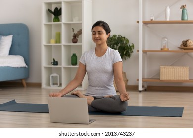 Calm Indian woman meditating, practicing yoga online at home, sitting on lotus pose on mat with laptop, breathing fresh air, stress relief, mindful young female involved in internet lesson, webinar