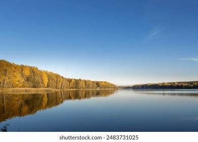 A calm lake with a blue sky in the background. The trees surrounding the lake are full of leaves, creating a beautiful autumn scene - Powered by Shutterstock