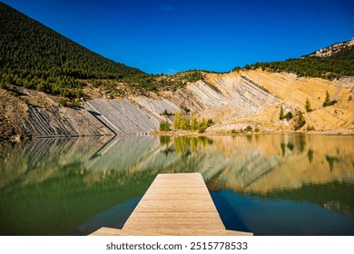 A calm lake reflects the surrounding hills and clear blue sky. A wooden pier extends into the water, creating a peaceful atmosphere. The rocky slopes and greenery along the shoreline enhance the scene - Powered by Shutterstock