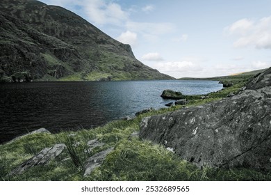 A calm lake rests between lush green hills, with rocky formations lining the shore. The sky is overcast, creating a peaceful atmosphere in a remote natural setting. - Powered by Shutterstock