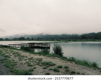 A calm lake scene with a wooden dock stretching out over the water, set against a backdrop of mist-covered mountains and an overcast sky, creating a serene and peaceful atmosphere. - Powered by Shutterstock