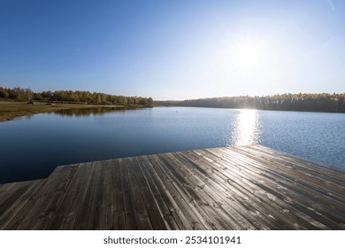 A calm lake with a wooden dock and a clear blue sky. The sun is shining on the water, creating a peaceful and serene atmosphere - Powered by Shutterstock