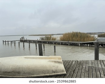 A calm lakeside scene on an overcast day, featuring a wooden dock extending over the water and a weathered rowboat resting on the shore. Reed plants add natural texture to the tranquil setting. - Powered by Shutterstock