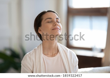 Calm leisure. Content young woman spending time at home alone, sitting on sofa with eyes closed in delight, breathing fresh cool air because of working conditioner, giving face to sunlight from window