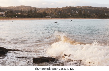 Calm Light Shines On A Wave In Foreground As It Breaks Over Rocks With Surfers In Background And A Beach Town In The Distance. Lorne, Victoria, Australia. Great Ocean Road.