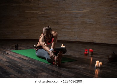 Calm Long Haired Sporty Woman Sitting On The Yoga Mat In Dark Room With Candles On The Floor And Stretching Her Body At The Yoga Practice