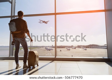 Calm male tourist is standing in airport and looking at aircraft flight through window. He is holding tickets and suitcase. Sunset