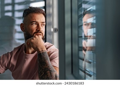 Calm man in pink t-shirt with tattoo on arms looking through window. - Powered by Shutterstock