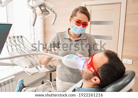 Calm man sitting in dentist chair while friendly female dentist in protective equipment adjusting apparatus for teeth whitening. Stomatologist working in dental clinic with male patient in the chair