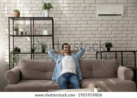 Calm man wearing glasses relaxing with closed eyes in room with air conditioner, sitting leaning back on cozy couch at home, breathing fresh air, satisfied customer enjoying comfortable temperature