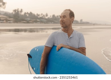 Calm mature man with bald head carrying blue surfboard walking along empty sandy beach at sunset and looking aside. Surfer in gray polo shirt holding surfing equipment spending vacation on shore. - Powered by Shutterstock